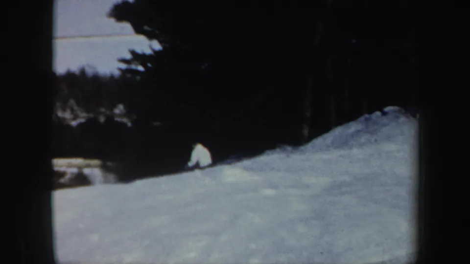1962 Vermont 8mm frame showing a distant skier on a snow-covered slope, bordered by dark trees and a pale open background.