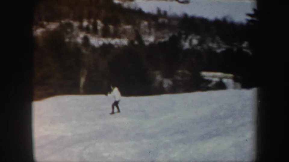 1962 Vermont home-movie frame of a lone skier on an open snowy slope, backed by wooded hills and a small structure or lift-like shape in the distance.