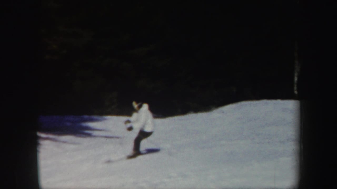 Skier in a white jacket moving across a snowy slope near dark trees.