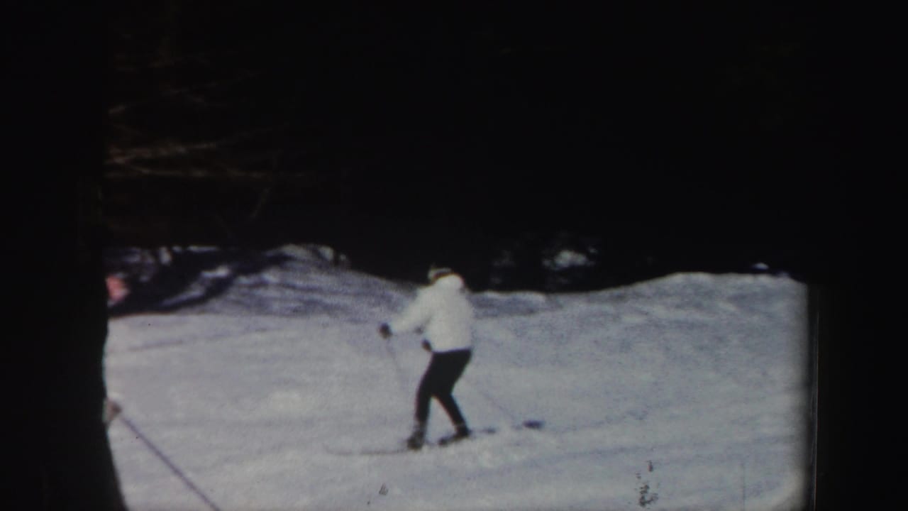 1962 Vermont home-movie frame of a skier in a white jacket and dark pants moving along a snowy slope before a wooded background.