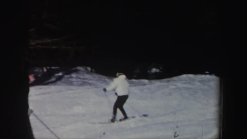 1962 Vermont home-movie frame of a skier in a white jacket and dark pants moving along a snowy slope before a wooded background.