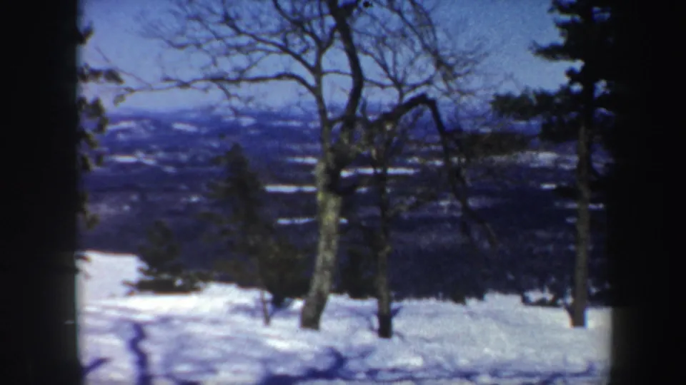 1962 Vermont home-movie frame with bare deciduous trees, snow cover, and a distant winter valley beyond the slope.