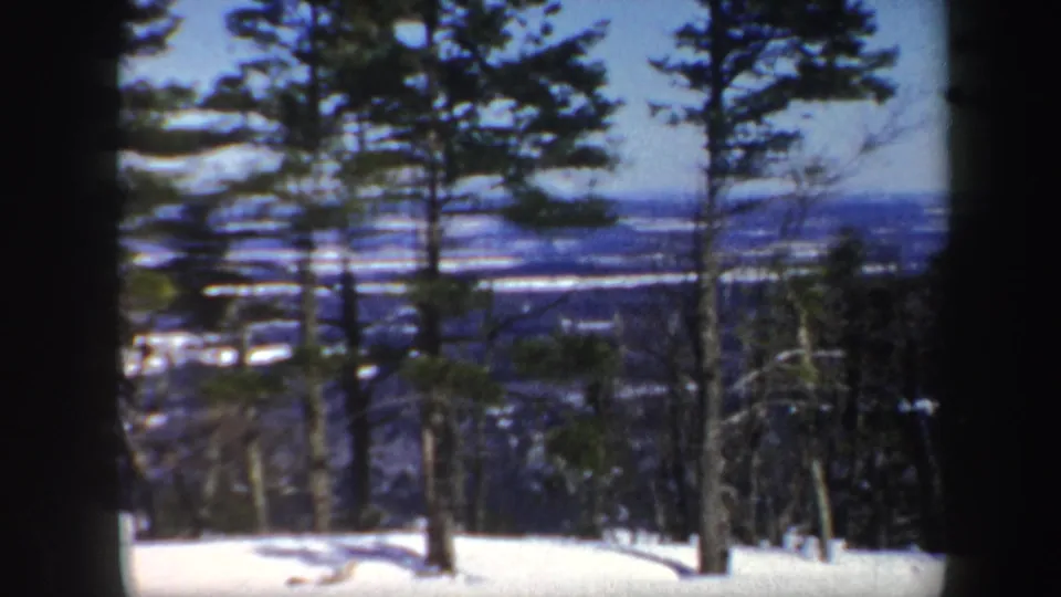 1962 Vermont 8mm frame showing snow-covered forest trees overlooking distant frozen water and low hills under blue sky.