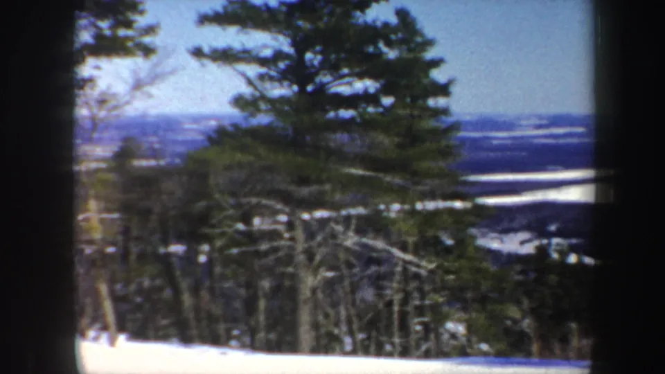 1962 Vermont 8mm frame looking past pines and winter woods toward a broad frozen lake or river in the valley.