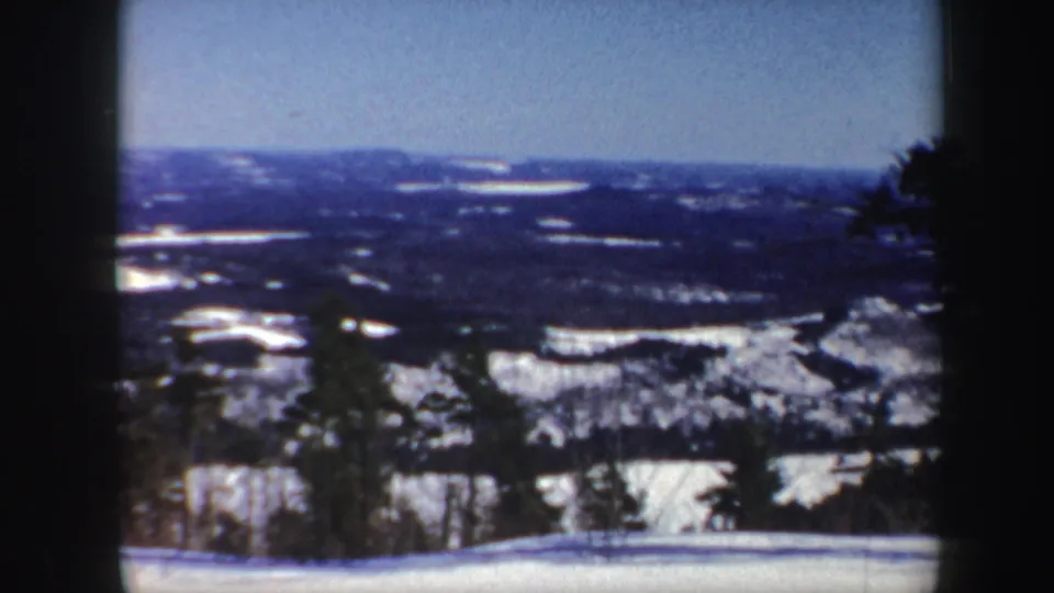 1962 Vermont home-movie frame with snow fields, distant wooded hills, blue sky, and dark foreground edges from the camera or film frame.
