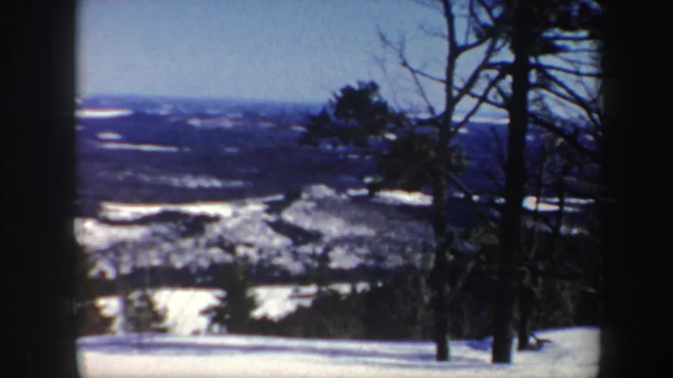 1962 Vermont 8mm frame showing a snowy slope, bare and evergreen trees, and a distant winter landscape below a pale blue sky.