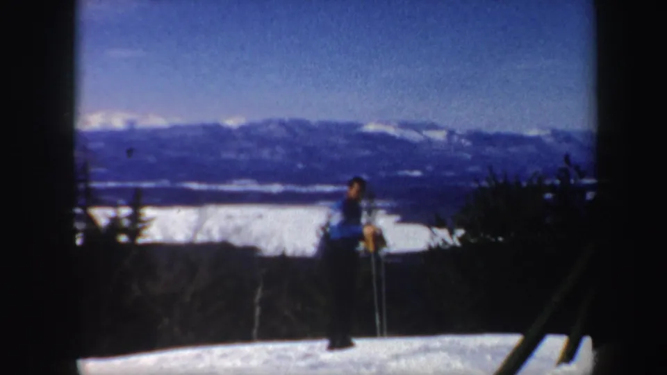 1962 Vermont home-movie frame with a distant person holding ski poles on a snowy ridge overlooking dark hills and bright winter sky.