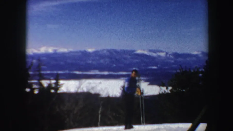 1962 Vermont 8mm frame of a lone skier with poles on a snowy ridge above a lake and distant winter mountains.