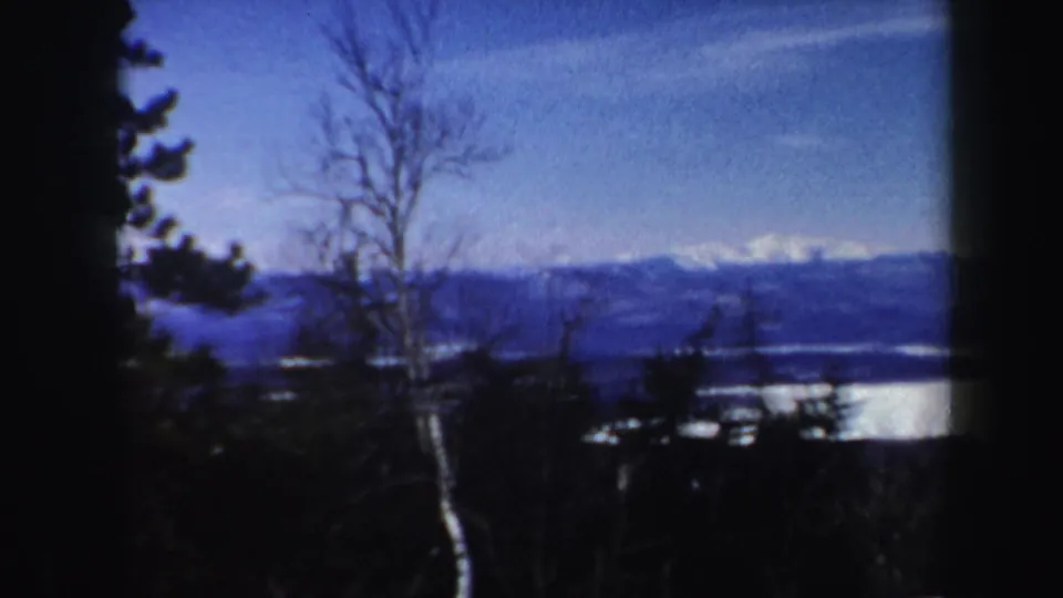 1962 Vermont winter landscape with bare trees, evergreens, a bright lake below, and snow-capped mountains in the distance.