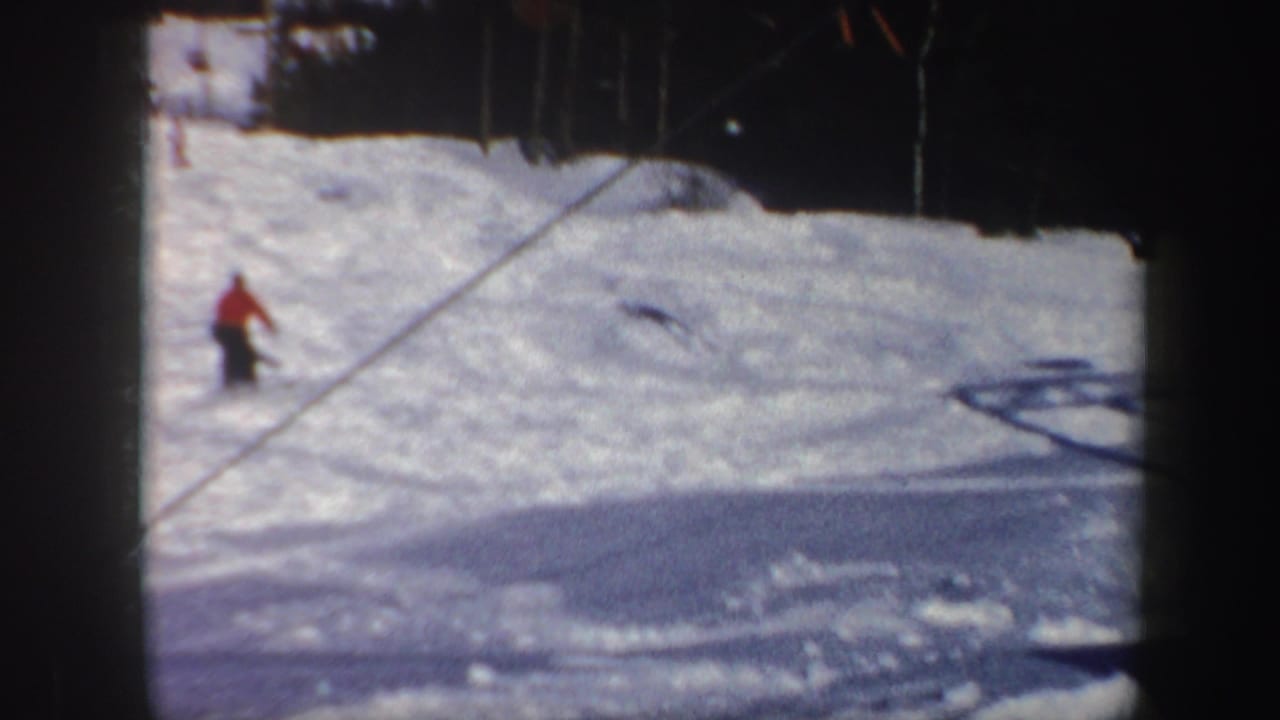 Skier in a red jacket descending a snowy slope with trees at the top edge.