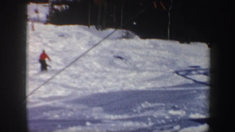 1962 Vermont 8mm frame showing a distant skier in a red jacket moving downhill across sunlit snow near a dark tree line.