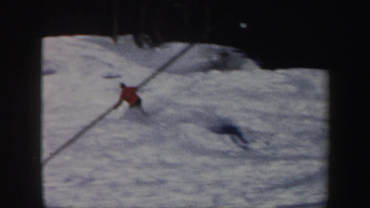 Red-jacketed skier leaning across the snowy slope with a long shadow behind them.