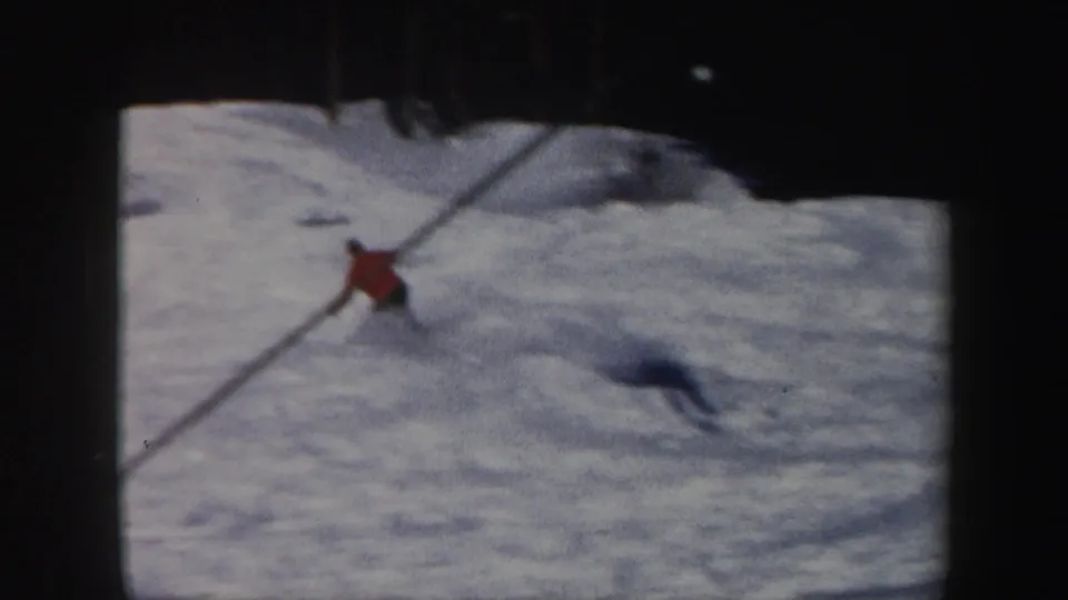 1962 Vermont 8mm frame of a red-jacketed skier carving across a bright snowfield with their shadow stretching over the slope.
