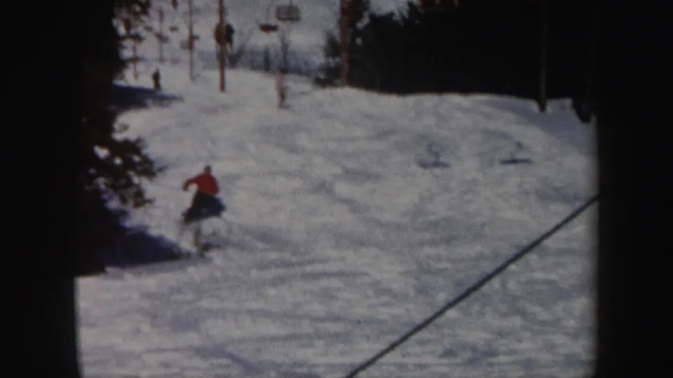 1962 Vermont home-movie frame showing a red-jacketed skier descending below lift poles on a tree-lined snowy ski slope.