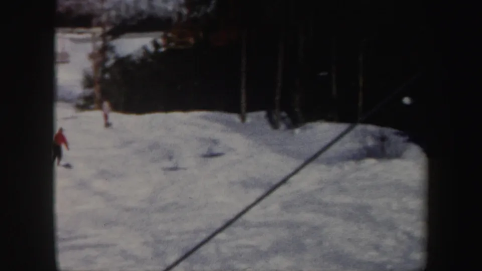 1962 Vermont 8mm frame with a distant red-jacketed skier crossing a sunlit slope near trees and ski-lift poles.