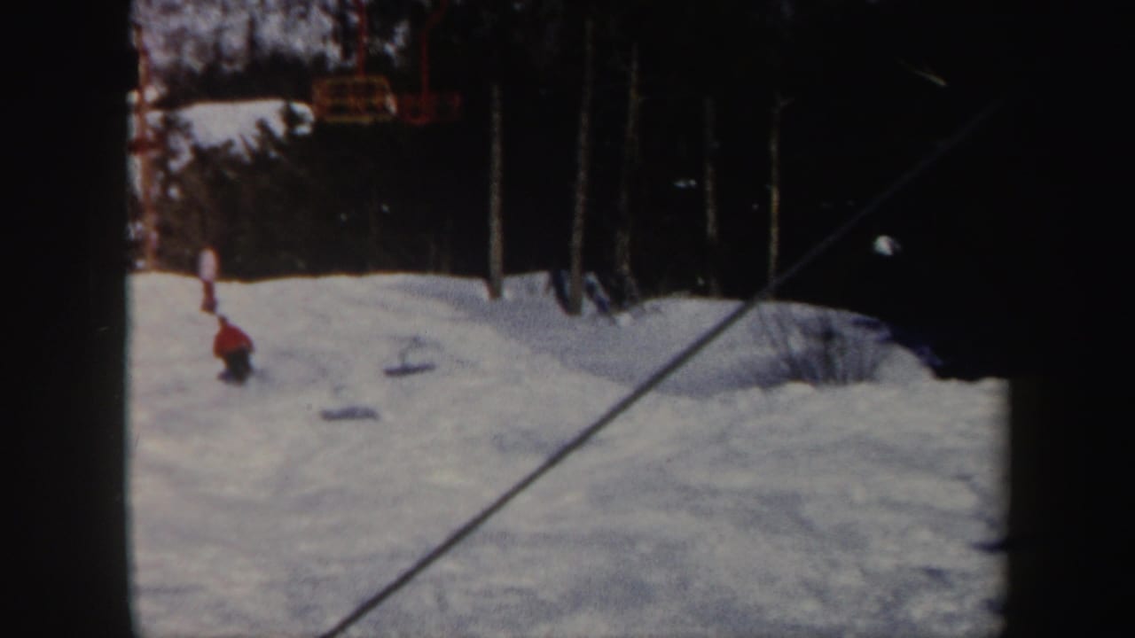 1962 Vermont home-movie frame showing a distant skier descending a snowy slope with lift cables and dark evergreens nearby.