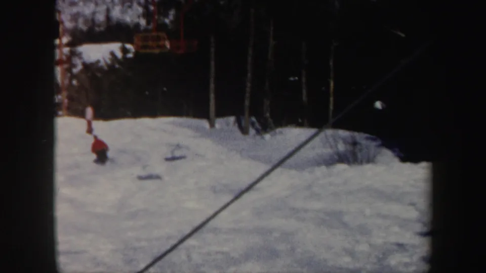 1962 Vermont home-movie frame showing a distant skier descending a snowy slope with lift cables and dark evergreens nearby.