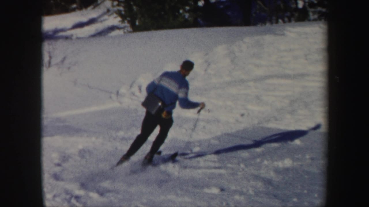 Skier in a blue sweater descends a snowy slope, casting a long shadow.