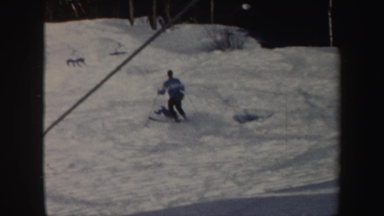 A skier in a light jacket moves across a snowy slope with lift cables and trees nearby.