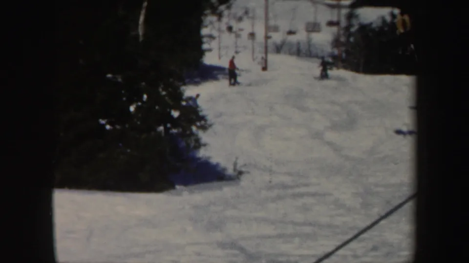1962 Vermont home-movie frame of a snowy ski run with small figures, lift equipment, and trees at the edge.