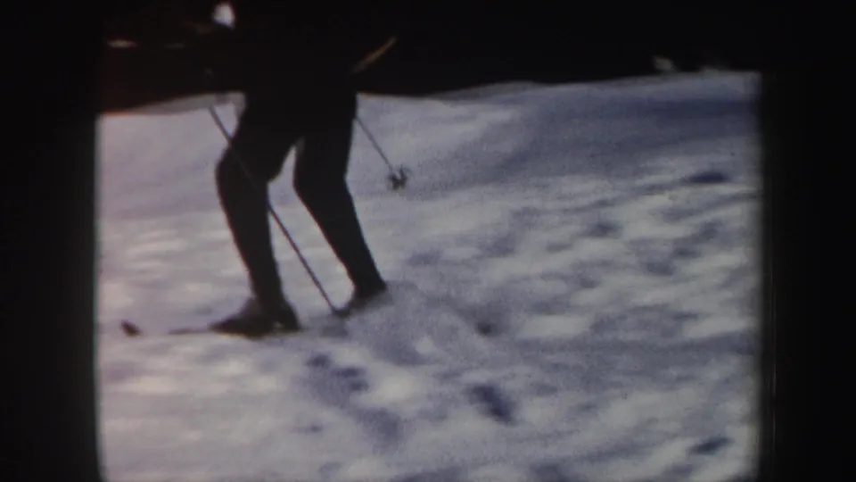 1962 Vermont home-movie frame showing a skier's lower body and poles crossing uneven snow on a resort slope.