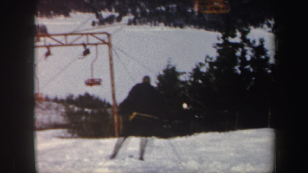 1962 Vermont ski resort frame with a skier moving downhill near a chair lift structure and dark evergreens.