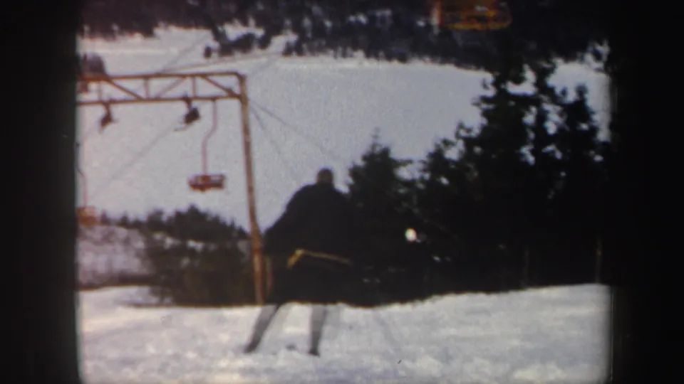 1962 Vermont ski resort frame with a skier moving downhill near a chair lift structure and dark evergreens.