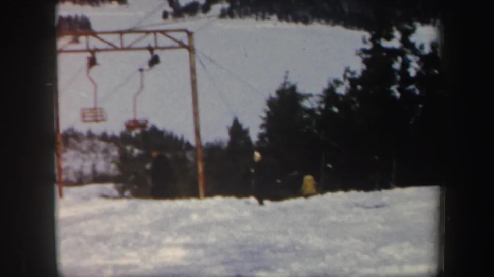 1962 Vermont resort frame with chair lift structures, suspended chairs, distant skiers, snow, and evergreens.