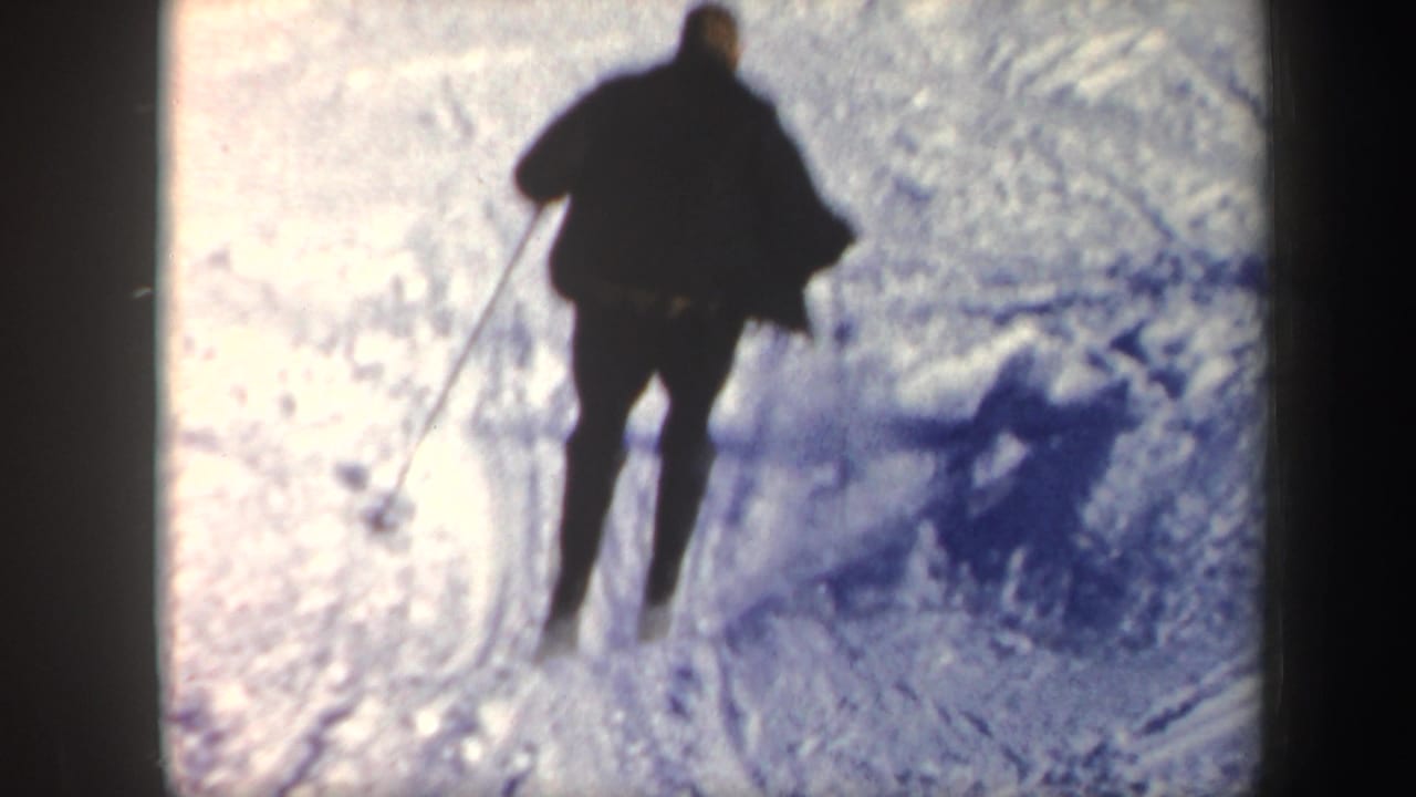 Skier in dark clothing moves across bright snow, seen from behind in soft 8mm focus.