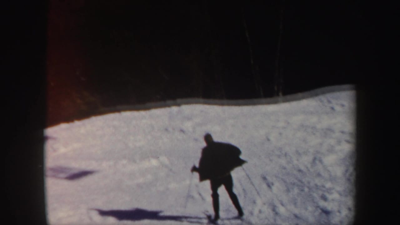 A distant skier in dark clothing traverses a bright Vermont snow slope, with trees at the ridge and a long shadow on the snow.