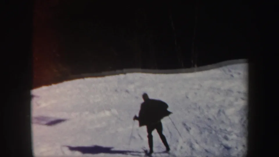 A distant skier in dark clothing traverses a bright Vermont snow slope, with trees at the ridge and a long shadow on the snow.