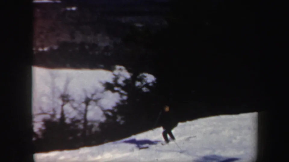 A skier is seen low on the snow-covered slope beneath bare winter trees, with the home-movie frame darkened at the edges.