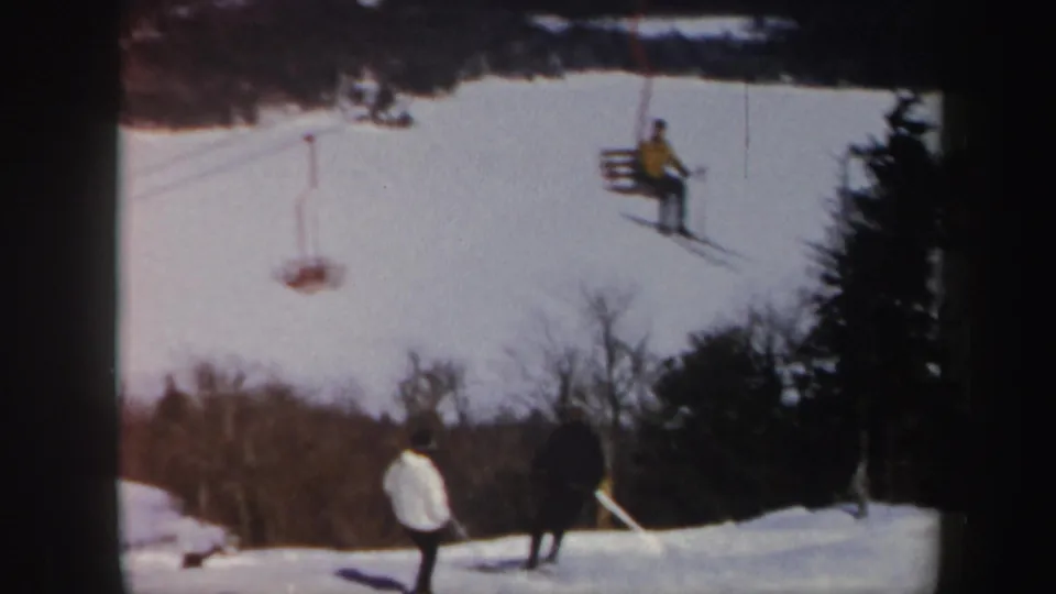 1962 Vermont ski-slope view with skiers on snow and a chairlift rider crossing the upper frame.