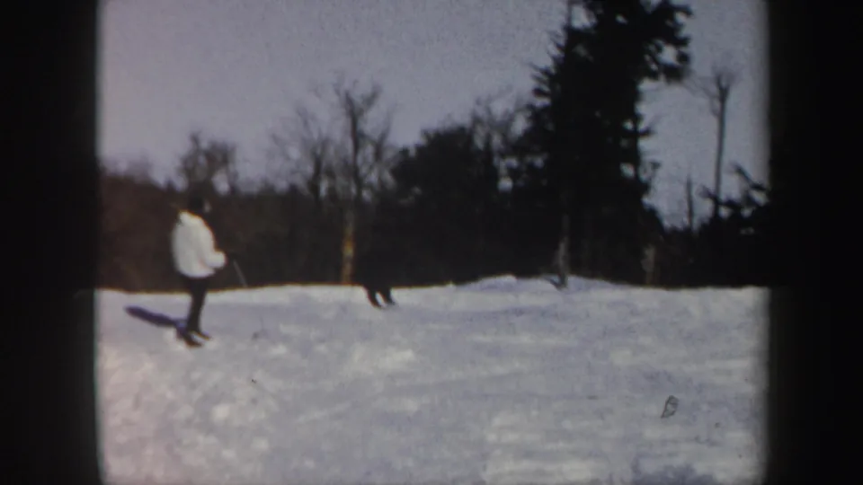 Two distant skiers traverse a Vermont ski area, framed by bare trees and dark evergreens in soft 8mm color.