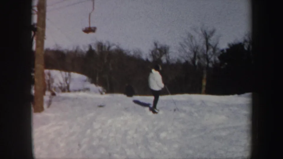 Distant skier in a light jacket on a snowy Vermont slope, with lift hardware, trees, and hillside snow behind.