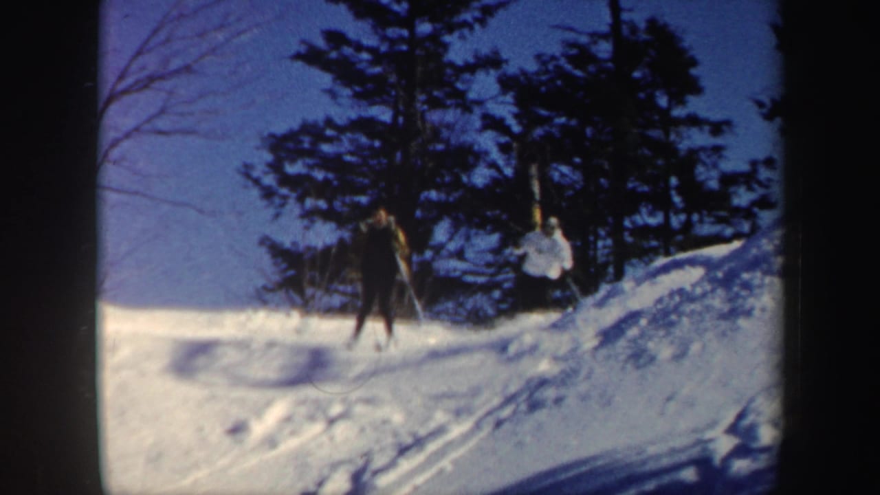 1962 Vermont home-movie frame showing two skiers crossing a snowy slope beneath bare trees and evergreens.