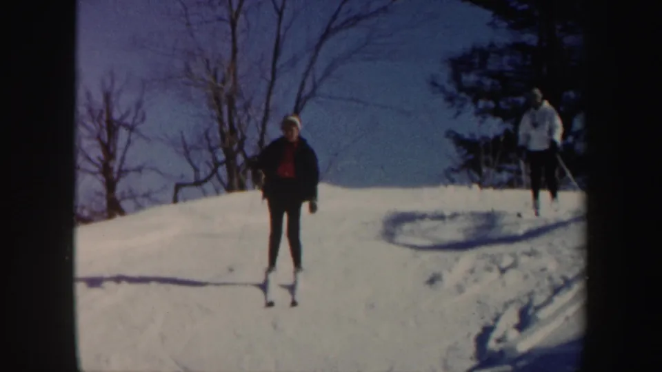 In 1962 Vermont home-movie footage, a woman leads a second skier down a sunny snow-covered hill bordered by trees.