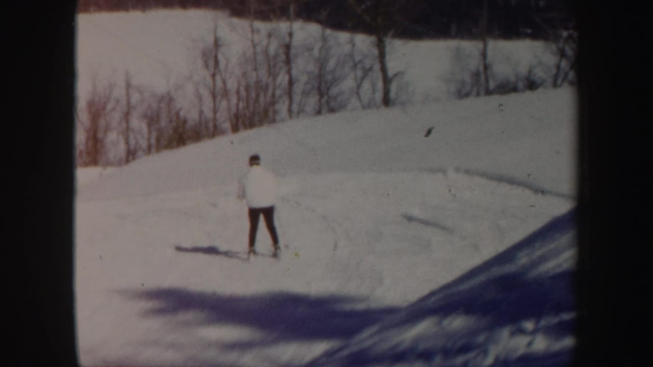 Skier in a white jacket crossing a snowy slope with bare trees behind.