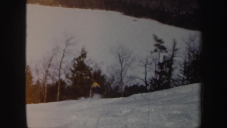 1962 Vermont home-movie frame with a small skier on a broad snowy slope and mixed winter trees beyond.