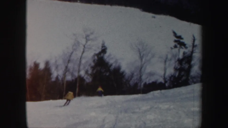 A skier in yellow moves downhill across a Vermont snow slope, with leafless trees and evergreens behind.