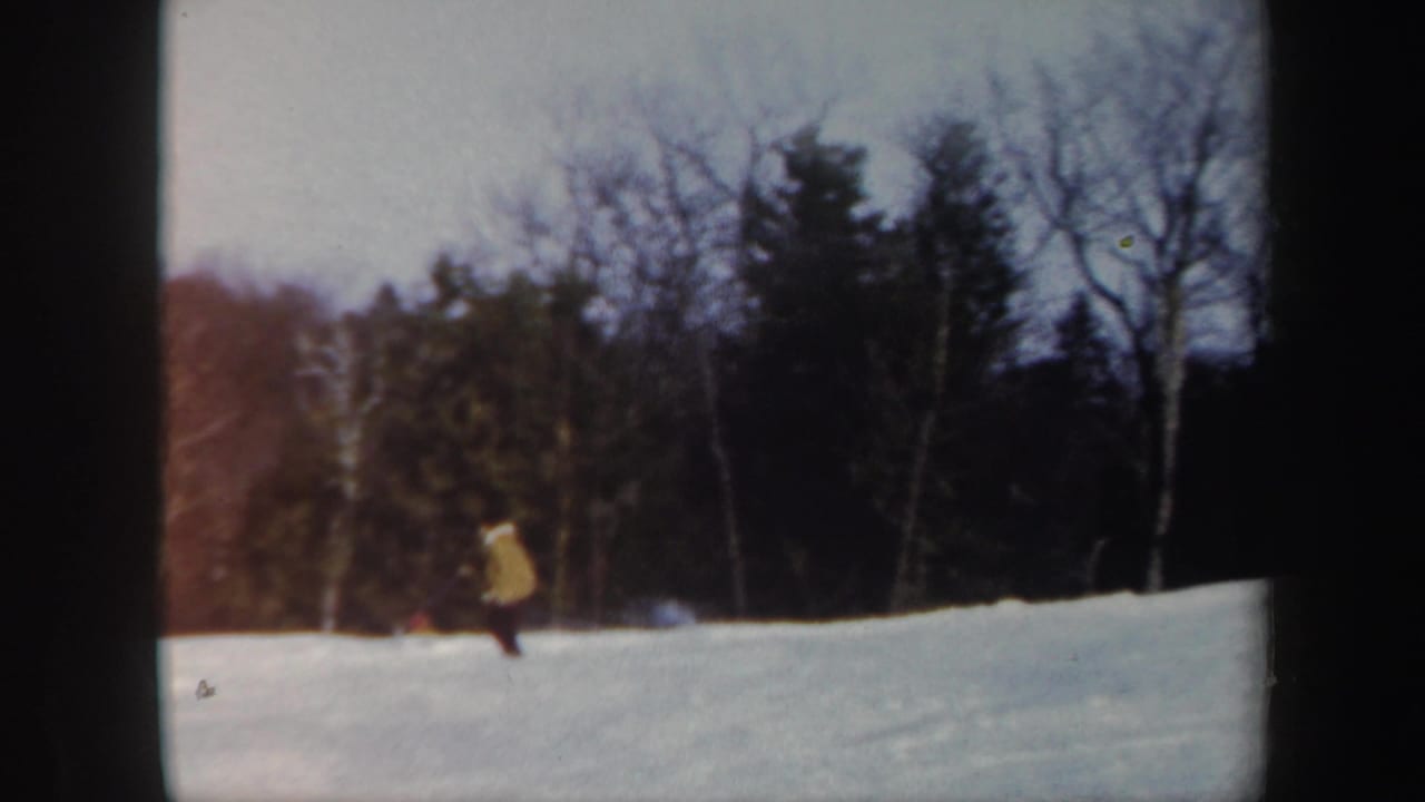 1962 Vermont 8mm frame showing a distant skier in a yellow jacket on a snowy slope with dark winter trees behind.