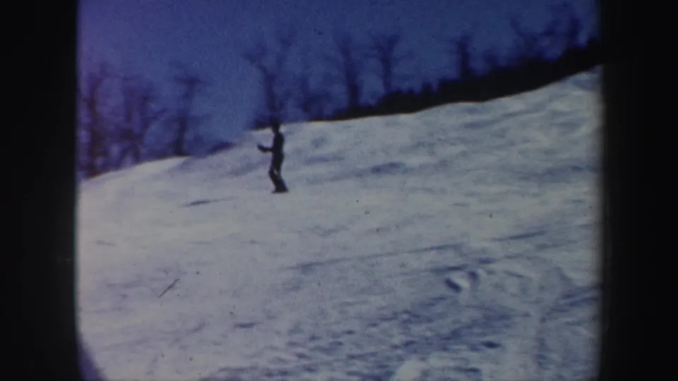 A distant skier, darker in exposure, crosses a blue-tinted snow slope with bare trees along the ridge in this 8mm frame.