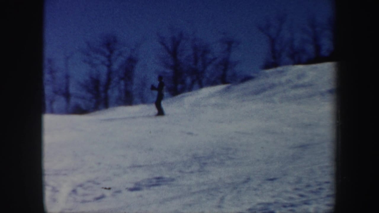 Distant skier on a snowy hillside beneath bare trees and blue sky.