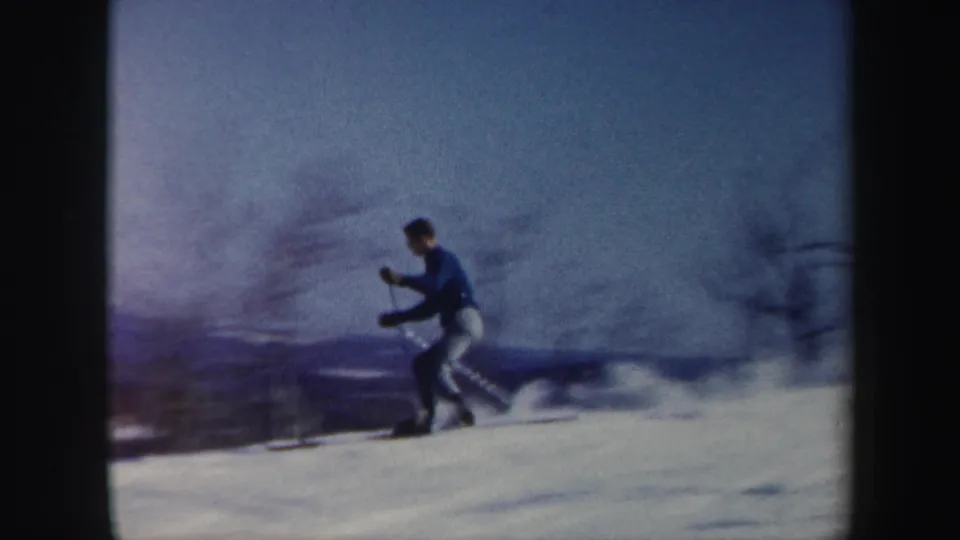 1962 Vermont 8mm frame of a young man skiing downhill, bent slightly forward with poles on a snow-covered slope.