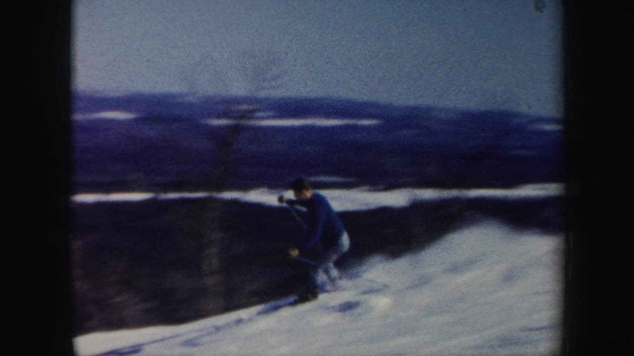 Skier in a blue sweater descends a snowy slope with distant hills behind.
