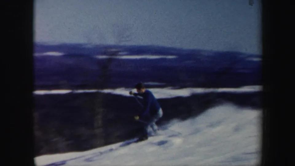 In Vermont 1962 home-movie footage, a skier in a blue sweater moves downhill across snow with dark hills in the background.