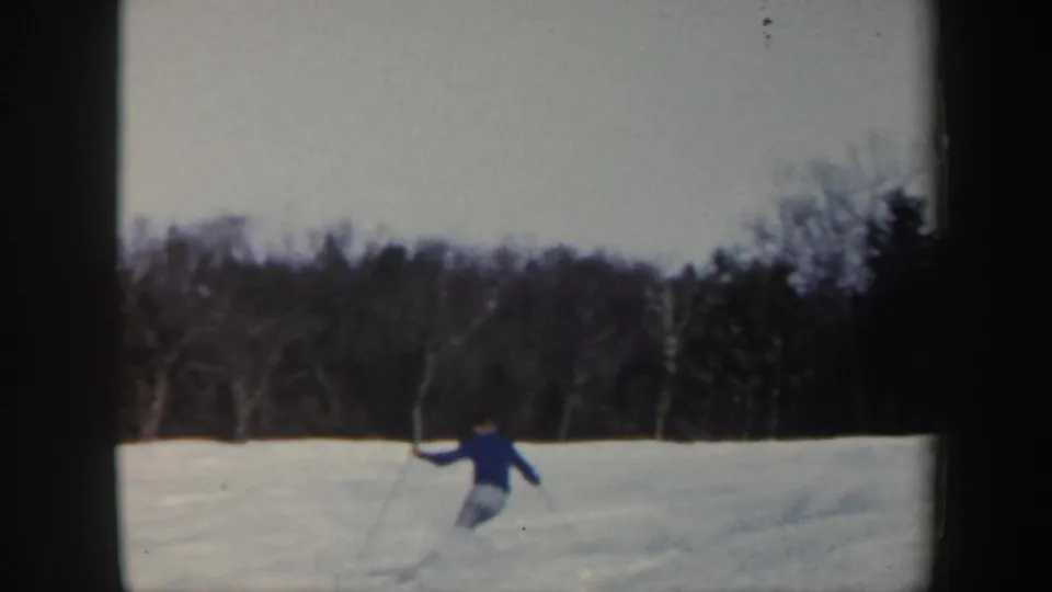 A distant skier in blue turns across an open snowy field in Vermont, framed by a dark winter tree line.