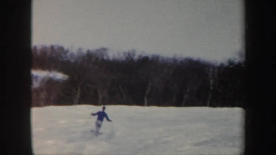 The skier appears small in the frame while descending a broad snowfield, with wooded Vermont terrain behind him.
