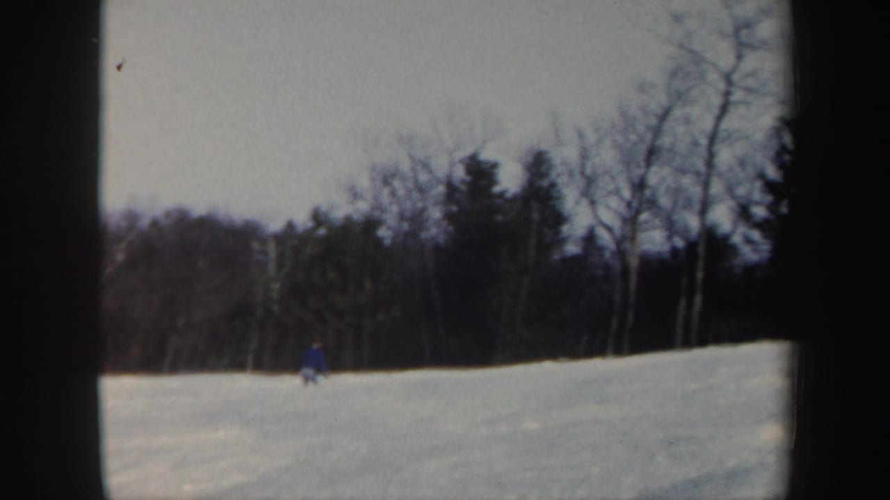 Small skier in blue descending a snowy hill below a tree line.