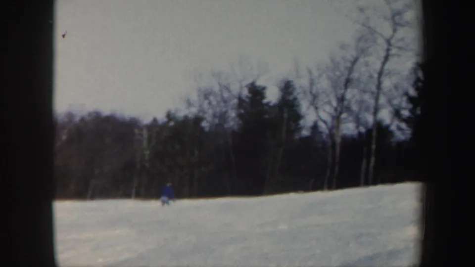 Distant 1962 Vermont skiing frame with a blue-clad figure on a gentle snow slope beneath bare winter trees.
